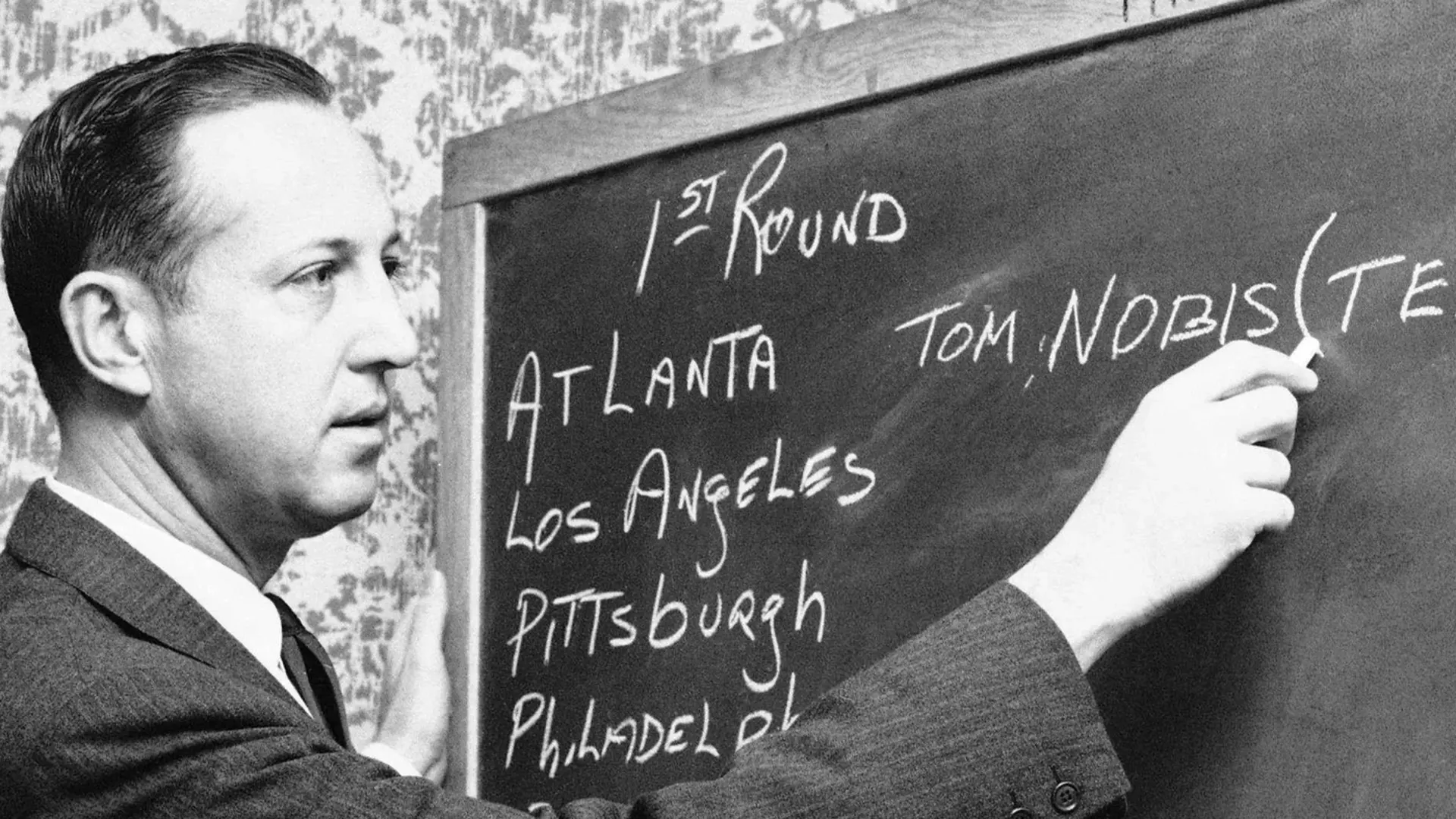 Black-and-white historical photograph of a man writing a first-round selection on an NFL draft board.